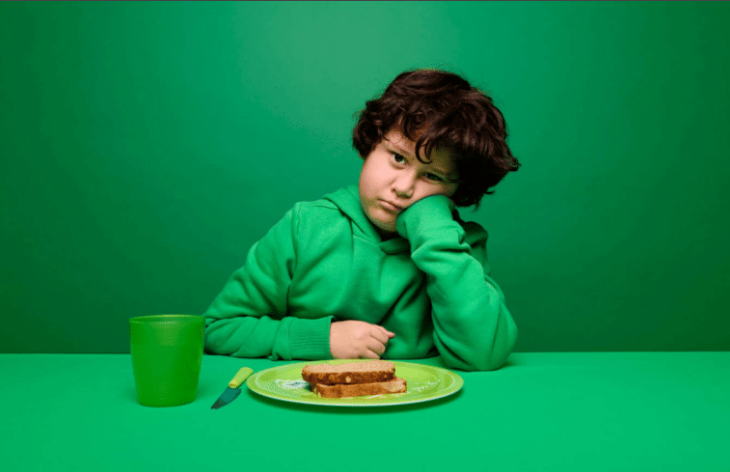 Kind zit met sip gezicht achter bord met boterham alsof hij er geen trek in heeft